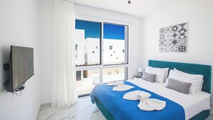 A hotel room with a large bed, a TV, and a window with a view of buildings. - Vie Bleu 1 - Four Bedroom Villa, Sleeps 8 (Πρωταράς)