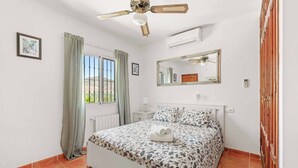 Ein Schlafzimmer mit Bett, Deckenventilator, Fenster mit Vorhängen, Nachttisch, Spiegel und Heizkörper.