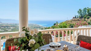 Ein Tisch mit Blumen in einer Vase, Tellern, Gläsern und Oliven auf einem Balkon mit Blick auf das Meer und die Hügel.