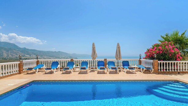 Ein Pool mit blauen Fliesen, Liegestühlen, Sonnenschirmen und Blick auf Berge.