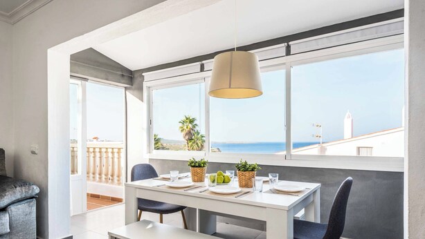 A dining area with a white table, two chairs, a bench, and a hanging light fixture. There are potted plants on the table and a view of the sea through large windows.