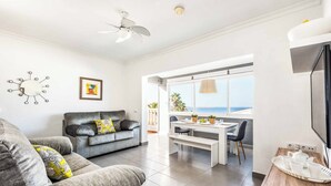 A modern living room with a grey sofa, a dining area with a white table, and a large window offering a view of the sea.