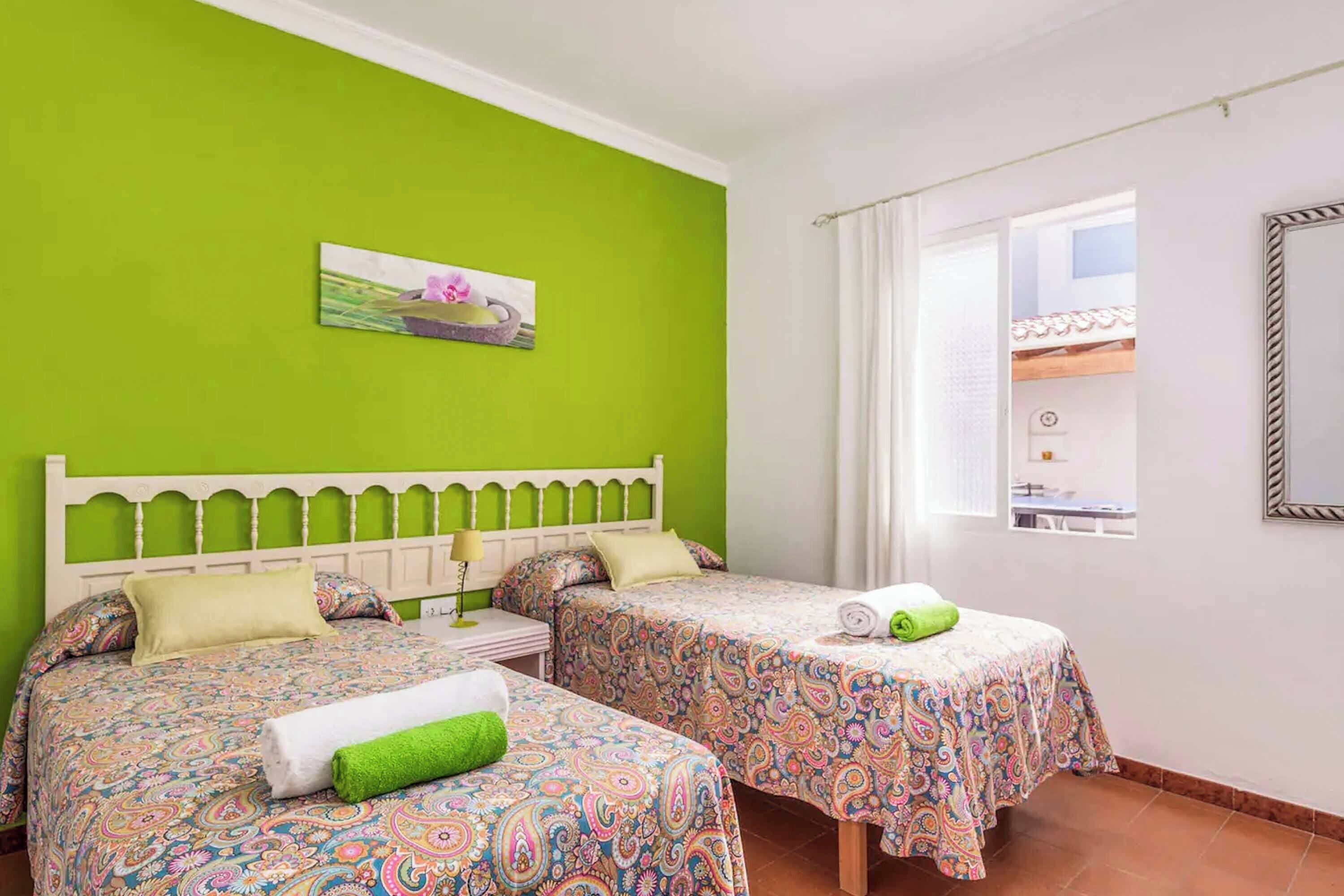 Une chambre avec deux lits, chacun recouvert d’une couvre-lit à motifs, un mur vert, un tableau et une fenêtre avec des rideaux.