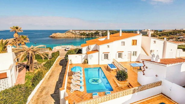 Una piscina en la azotea con sillones, vistas al mar y edificios blancos con tejados anaranjados.