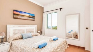 Un dormitorio con una cama grande, una mesita de noche con lámpara, una ventana con vistas y un cuadro en la pared.