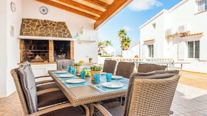 Un espacio de comedor al aire libre con mobiliario de mimbre, una chimenea de piedra y vistas a edificios blancos y palmeras.