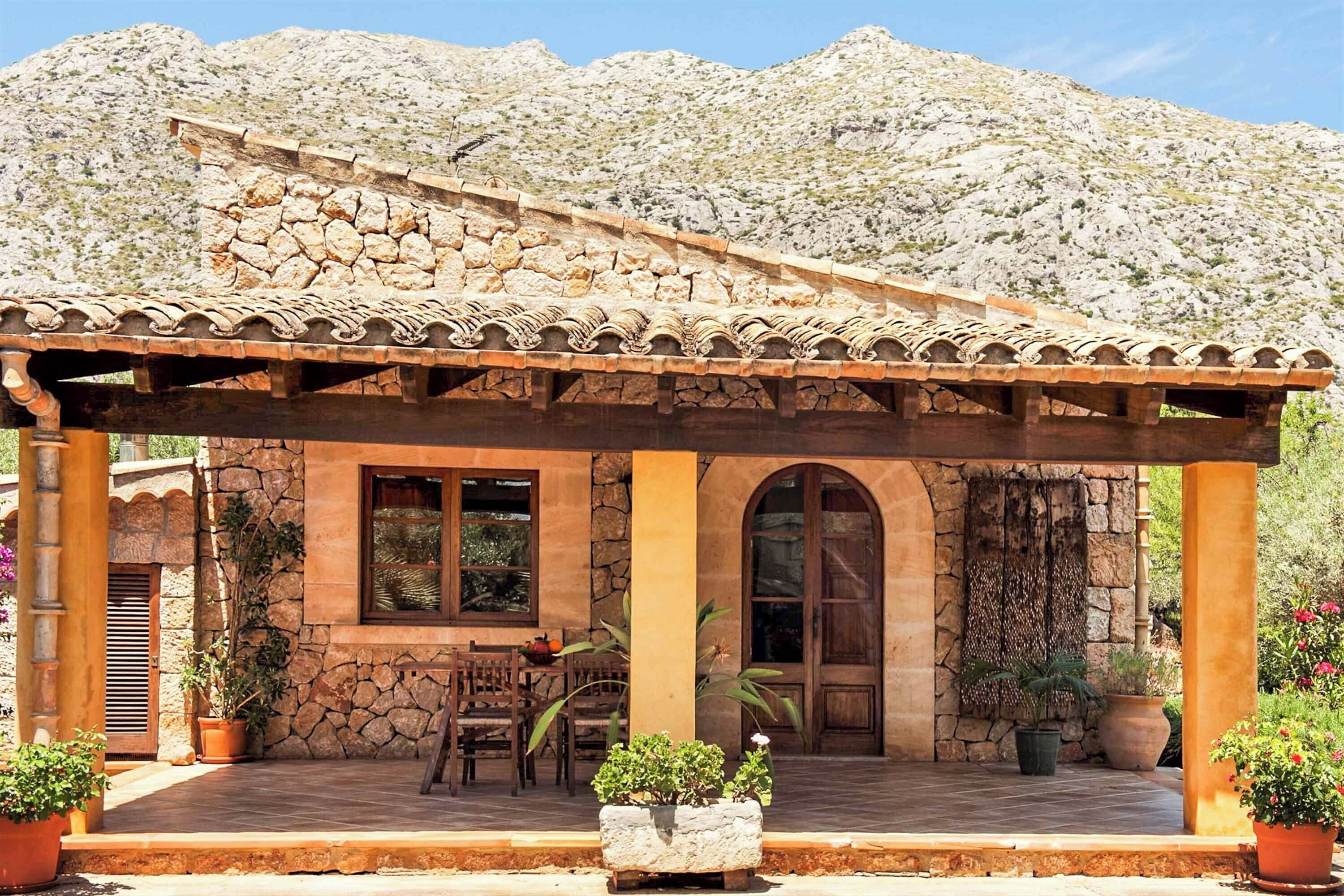 A stone building with a tiled roof, a covered patio with chairs, and potted plants.