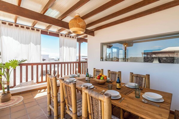 A dining area with a wooden table set for six, surrounded by bamboo chairs, a fruit bowl, and a bottle of wine.