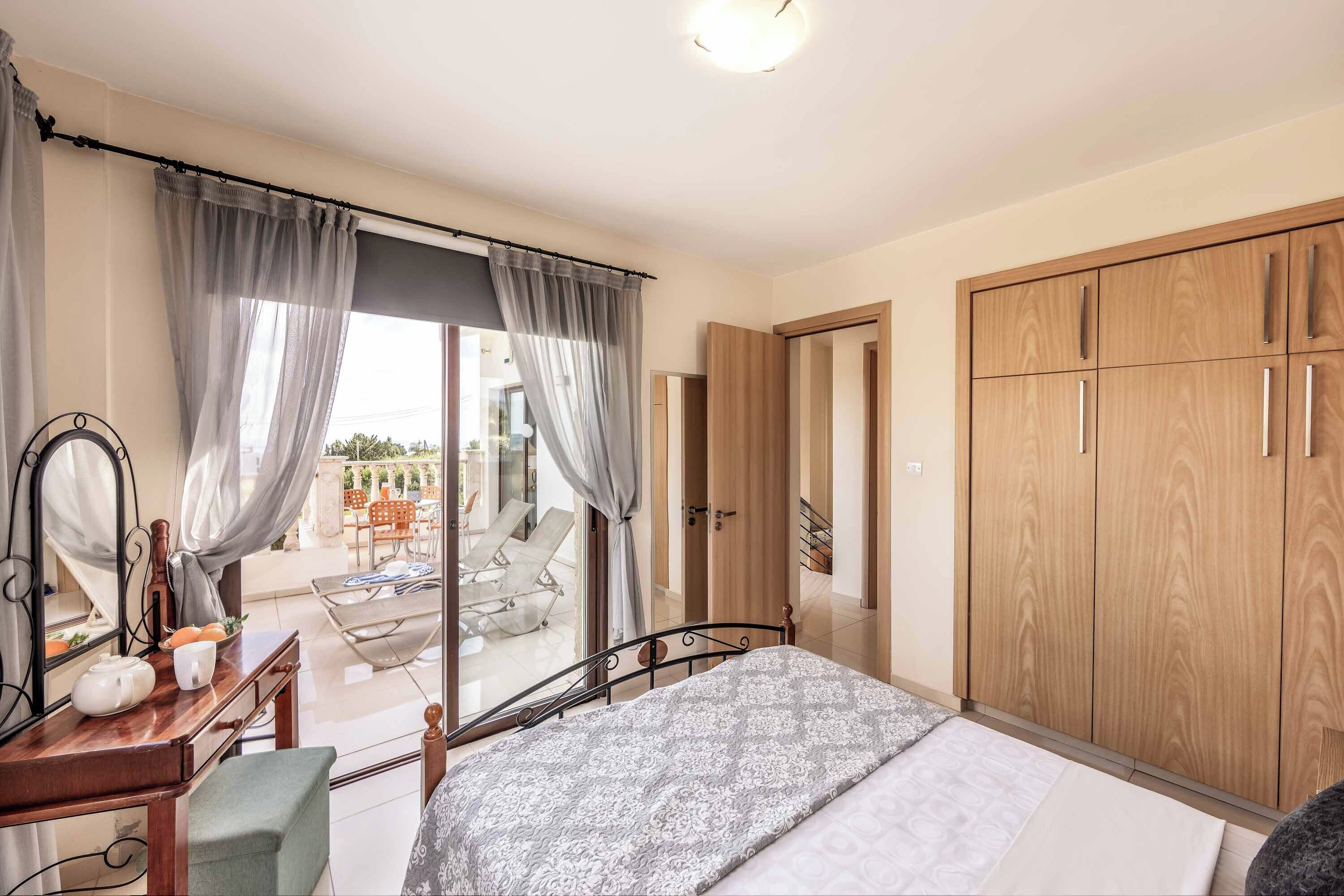 Een slaapkamer met een bed, een bureau, een spiegel, een kledingkast en een schuifdeur die leidt naar een buitenruimte met een tafel en stoelen.