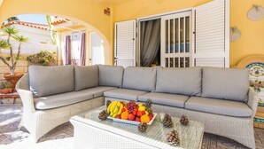 A wicker sofa with grey cushions, a glass coffee table with fruit and pinecones, and a white door with shutters.