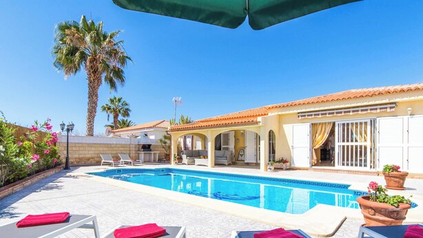 A pool area with lounge chairs and a pool, surrounded by palm trees and a building with a tiled roof.