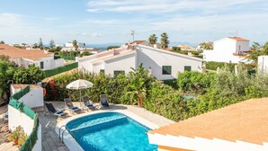 Una piscina en la azotea con sillones de descanso, rodeada de vegetación y edificios blancos con tejados rojos.