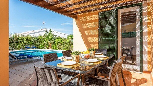 Un área de comedor al aire libre con piscina, sillas de mimbre y una mesa preparada para dos.