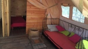 Interior de una cabaña de madera con una cama, una mesita y un ventanal con cortinas.