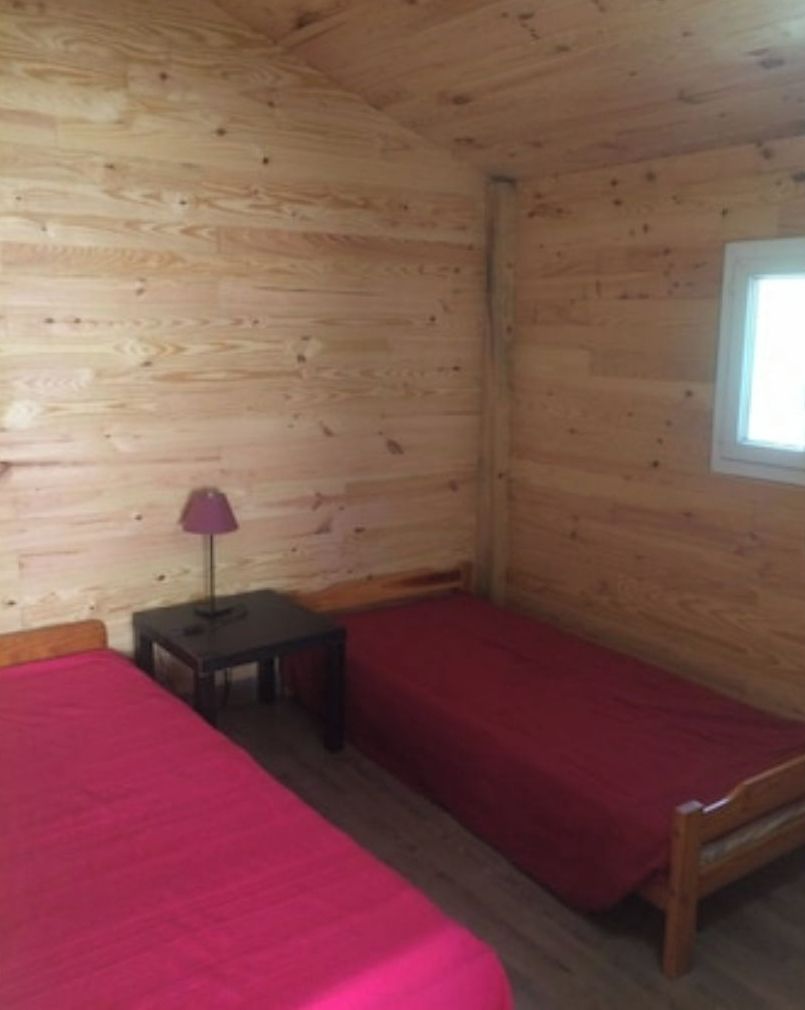 A room with wooden walls and a window. There are two beds with red bedding and a small table with a lamp.