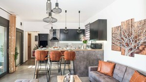 Eine moderne Küche mit Pendelleuchten, ein Essbereich mit Tisch und Stühlen sowie ein Wohnzimmer mit Sofa und Kissen.