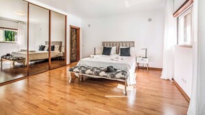 Een ruime slaapkamer met een groot bed, een bankje, een nachtkastje, een spiegel en een raam.