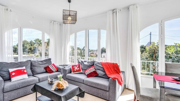 A modern living room with a grey sofa set, a coffee table, and a dining area with a red table runner. Large windows provide a view of the outdoors. - Villa Benicuco - Twelve Bedroom Villa, Sleeps 24 (Calp)