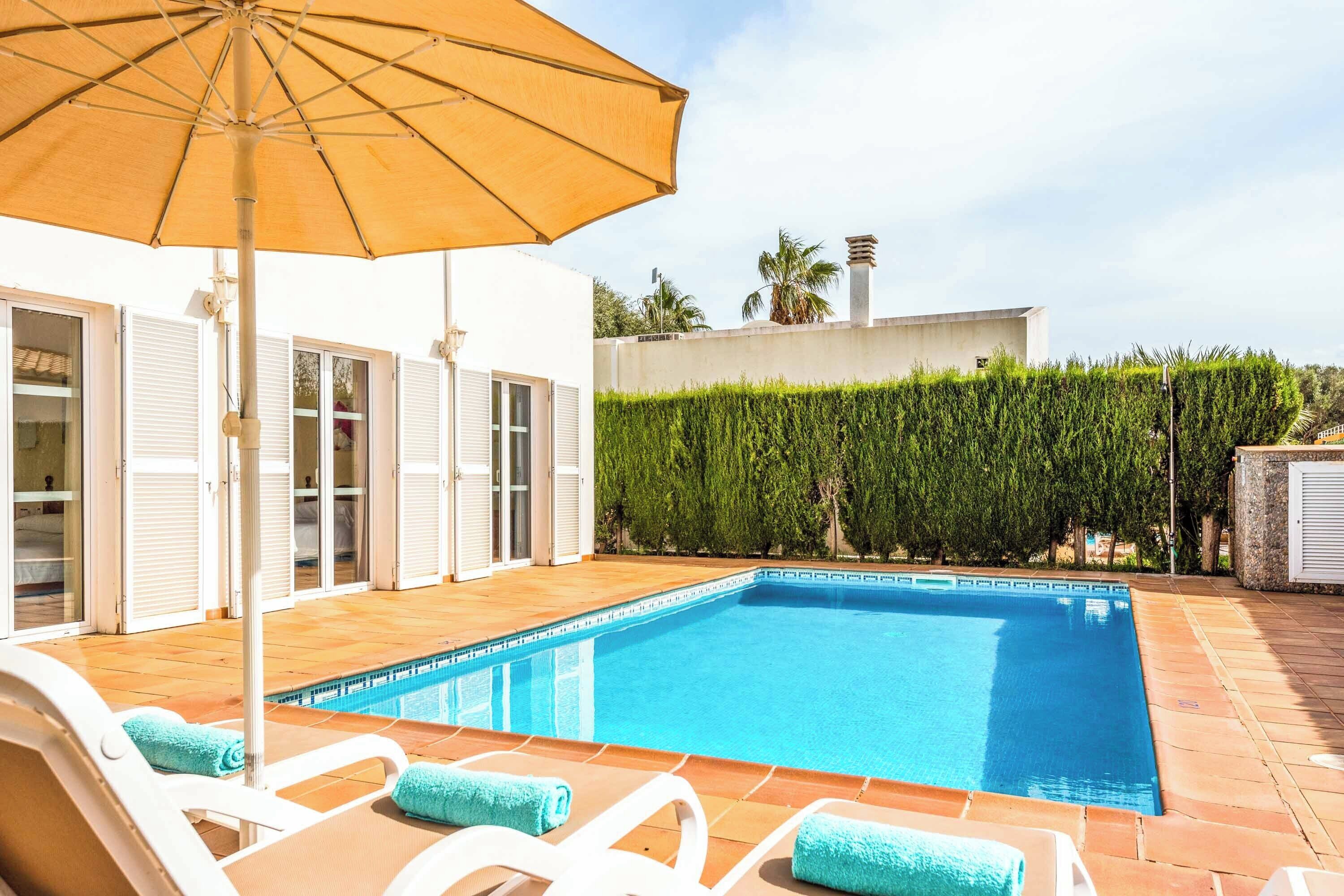 Una piscina con sillas de descanso y una sombrilla, rodeada por un edificio blanco con puertas de cristal y un seto.
