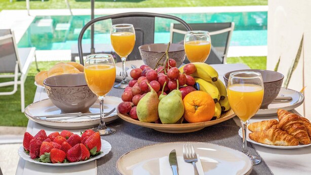 Une table de petit-déjeuner avec des fruits, des croissants et du jus d’orange, placée au bord d’une piscine.