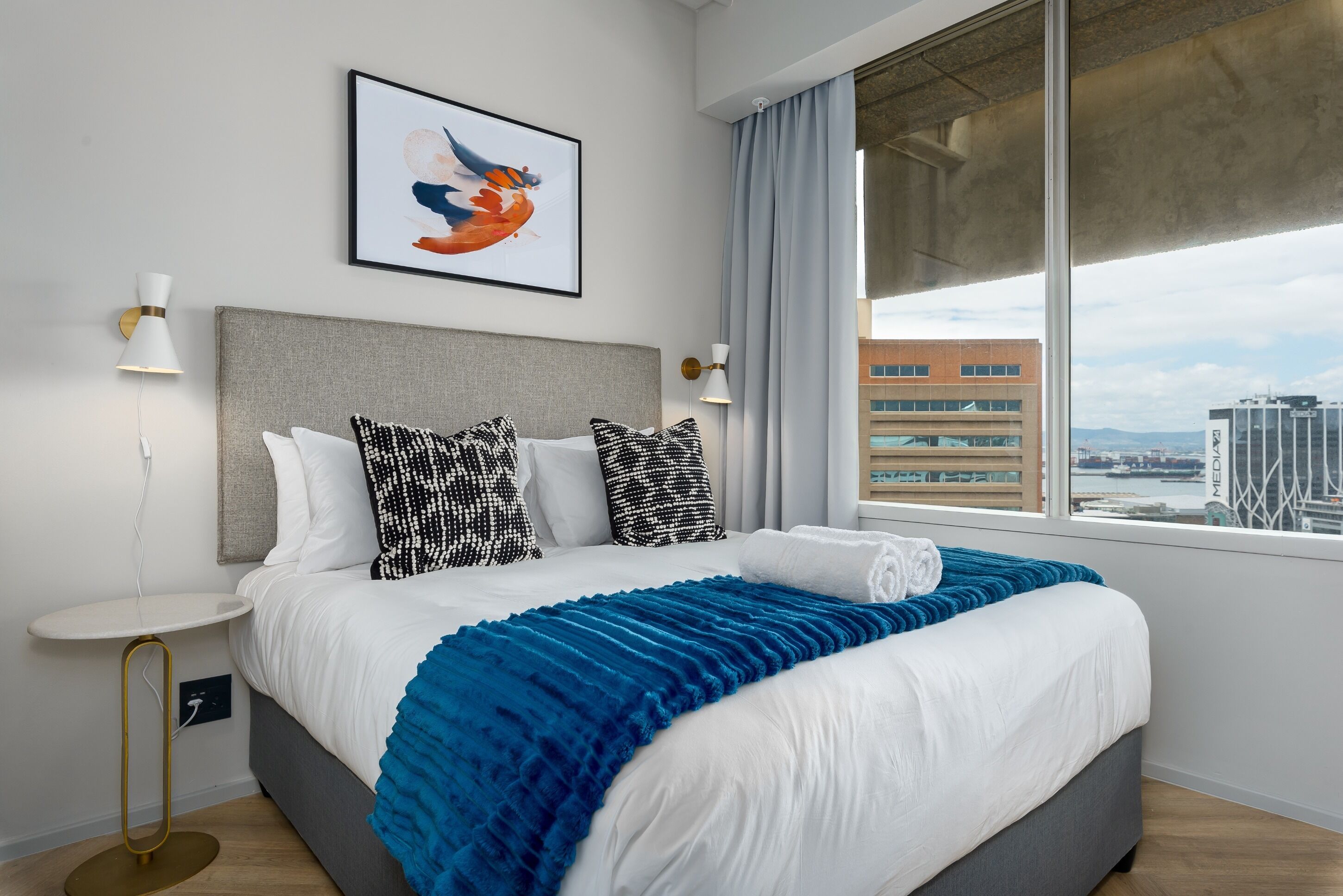 Una habitación de hotel moderna con una cama grande, vistas de la ciudad y una obra enmarcada en la pared.
