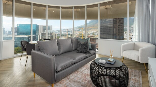 Ein modernes Wohnzimmer mit einem grauen Sofa, einem weißen Sessel und einem runden schwarzen Couchtisch. Große Fenster bieten Blick auf die Stadt.