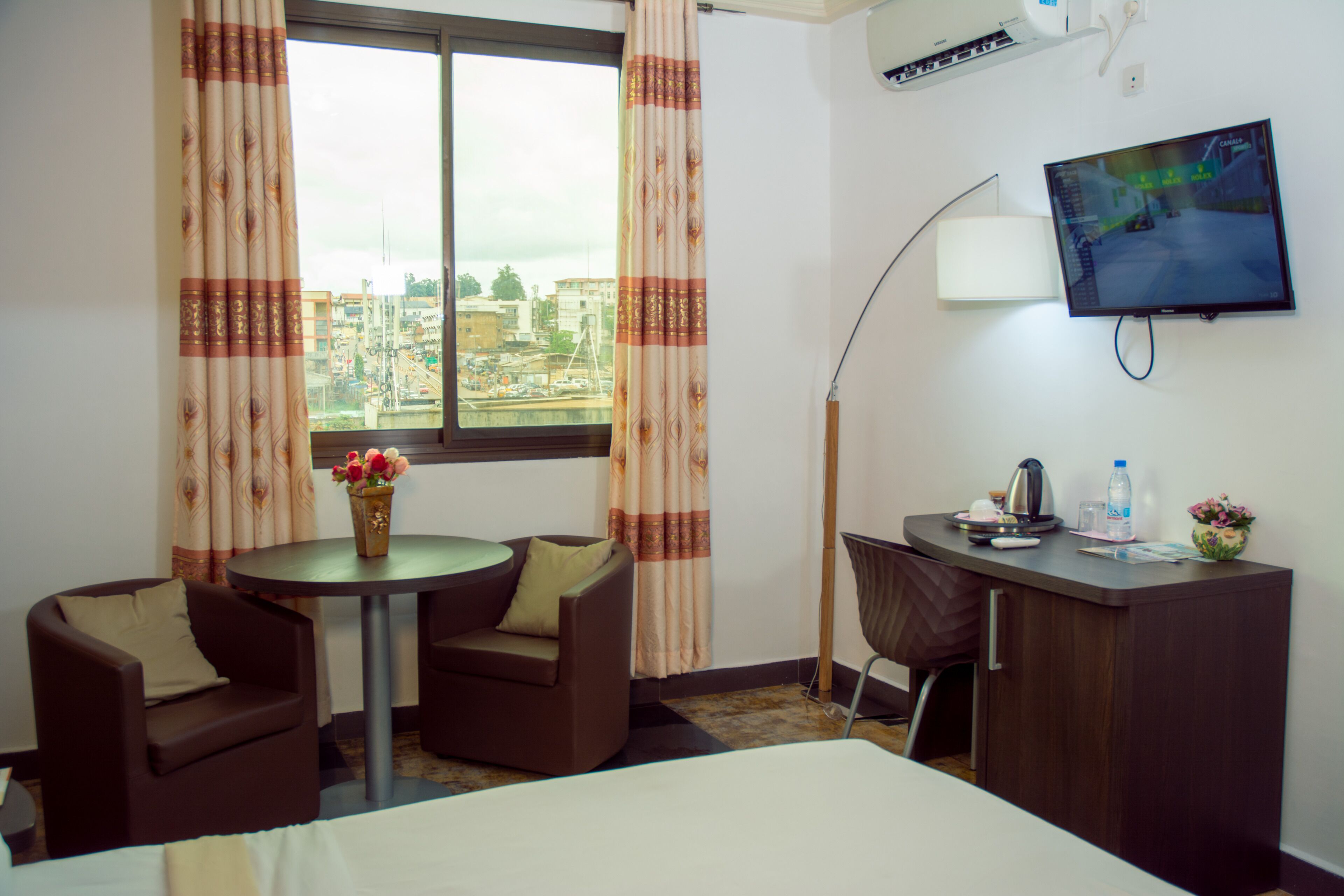 Een hotelkamer met een bed, twee fauteuils, een bijzettafeltje met een bloevas, een bureau met een computer, een lamp en een televisie aan de muur.