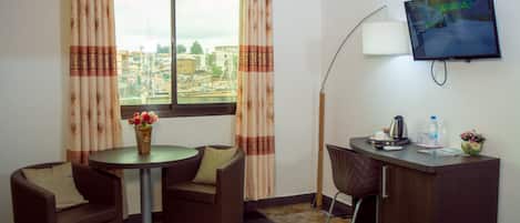 A hotel room with a bed, two armchairs, a small table with a vase of flowers, a desk with a computer, a lamp, and a TV on the wall.