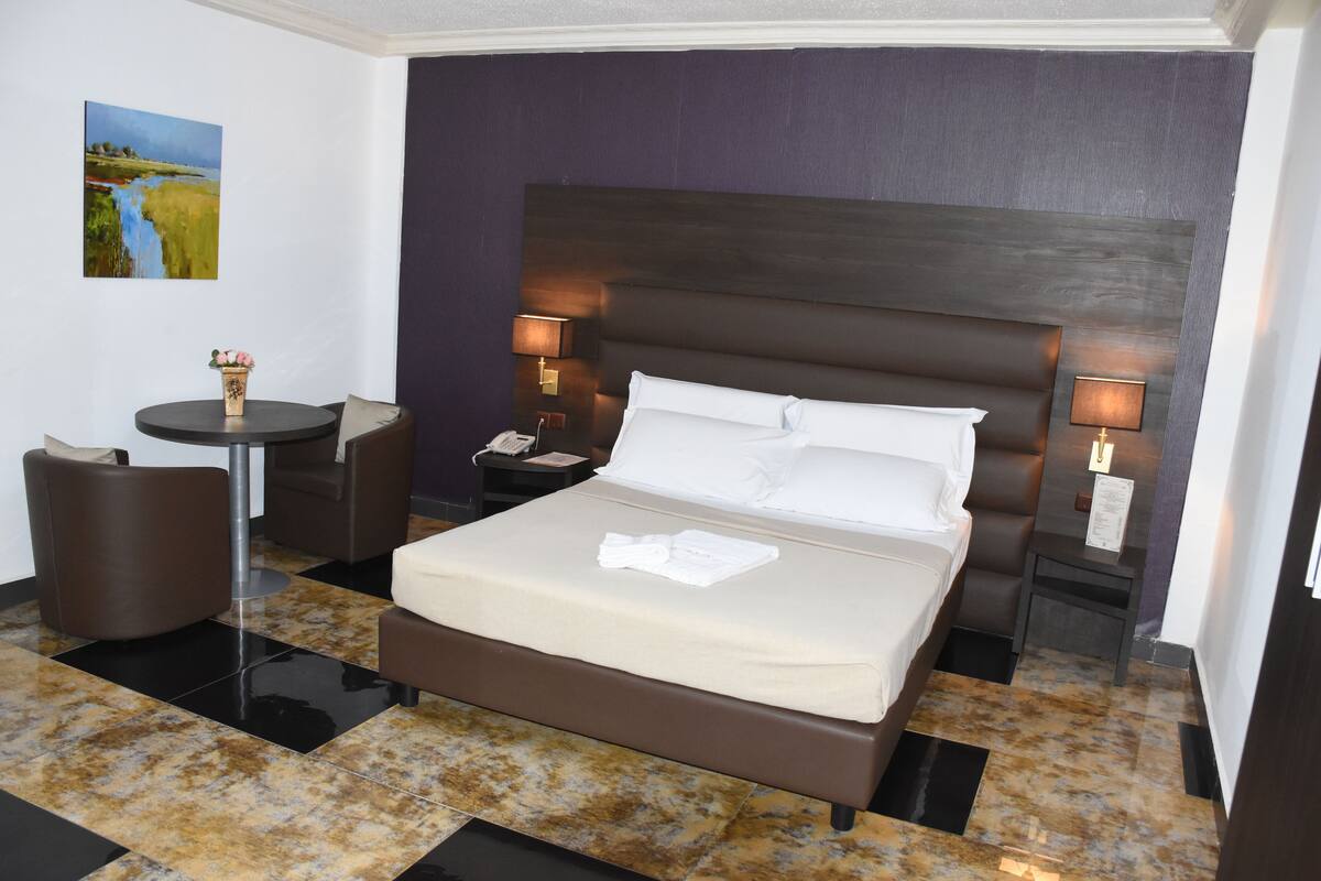 A hotel room with a large bed, a small round table, a chair, and a nightstand with a lamp.