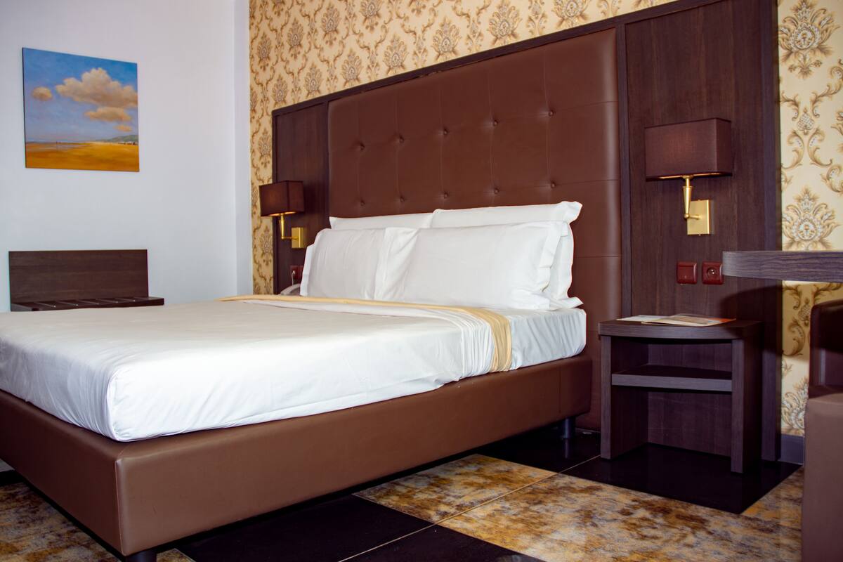 A neatly made bed with a brown headboard, a bedside table with a lamp, and a wall with a patterned wallpaper and a painting of a beach.