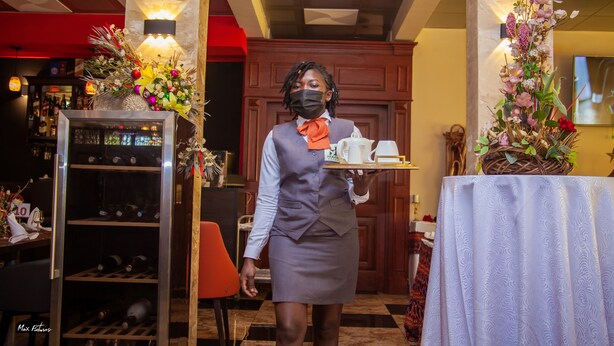 A hotel staff member carrying a tray with a teapot and cups, standing in a room with a wine rack and decorative flowers. - New Heaven Hôtel (Yaoundé)