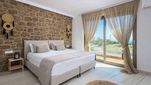 A bedroom with a stone wall, a large bed, a nightstand, and a view of the outdoors. - Villa Elios Residence (Rhodes)