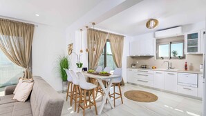 A modern kitchen with white cabinets, a dining area with a white table and chairs, and a living area with a sofa and curtains. - Villa Elios Residence (Rhodes)