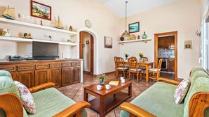 A cozy living room with a wooden coffee table, two green sofas, a TV, and a dining area with wooden chairs and a table.