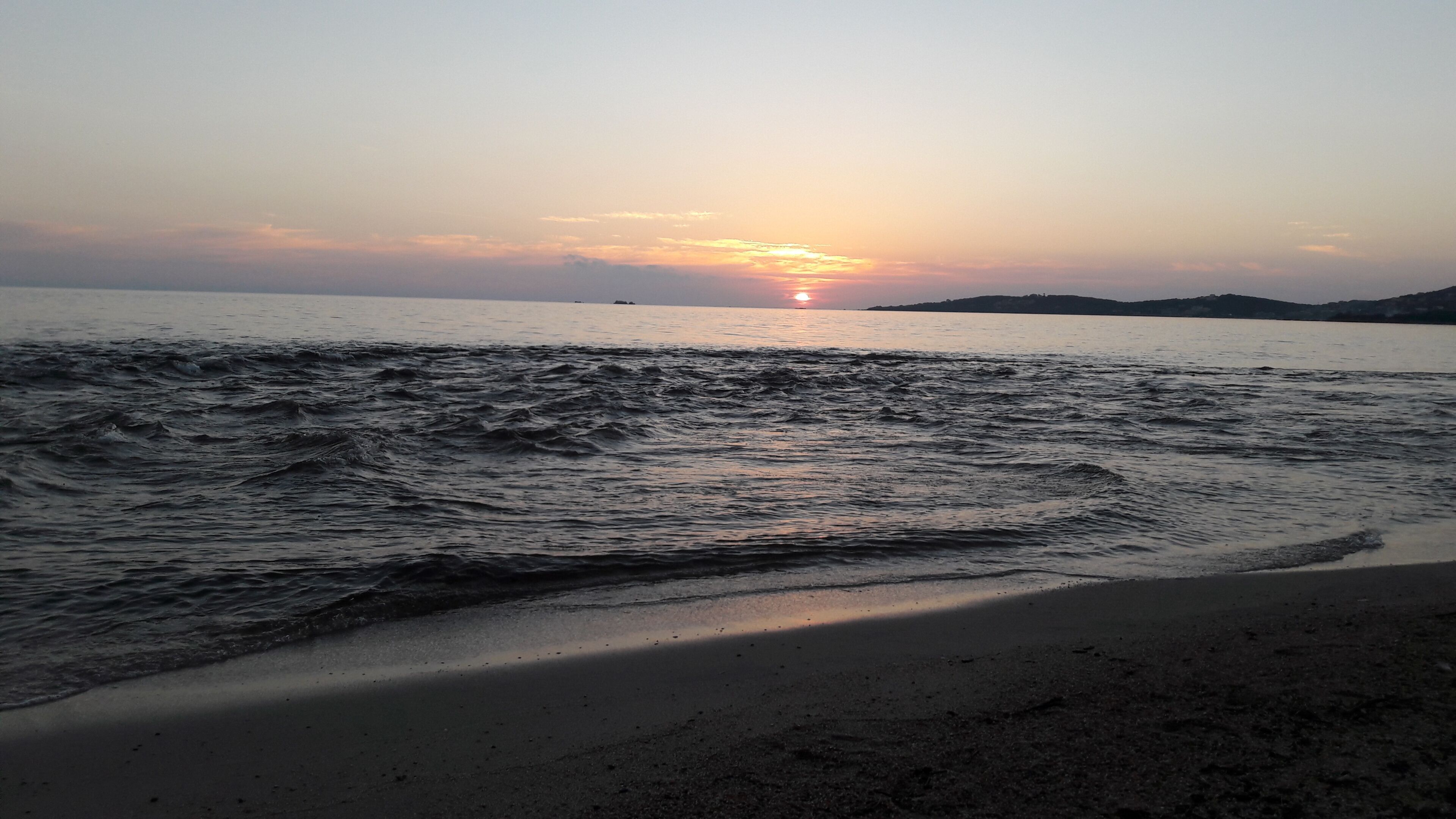 Een strand met golven, een zonsondergang en land in de verte.