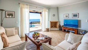 A living room with a sofa, coffee table, and TV. There is a view of the sea through a glass door. - Villa Tramonto Minies (Ionian Islands)