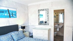 Un dormitorio con cama, mesita de noche, cómoda, espejo y baño visible a través de una puerta abierta.