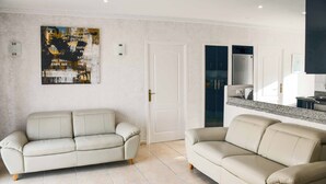 Una sala de estar moderna con dos sofás beige, una puerta blanca y una zona de cocina con encimera y armarios.