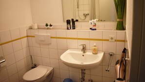 Une salle de bain avec des toilettes, un lavabo et une douche. On trouve des articles de toilette sur le comptoir et une plante devant le miroir.