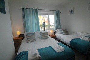 A hotel room with two beds, each with a blue bedspread and white linens, a window with curtains, and a view of a building outside.