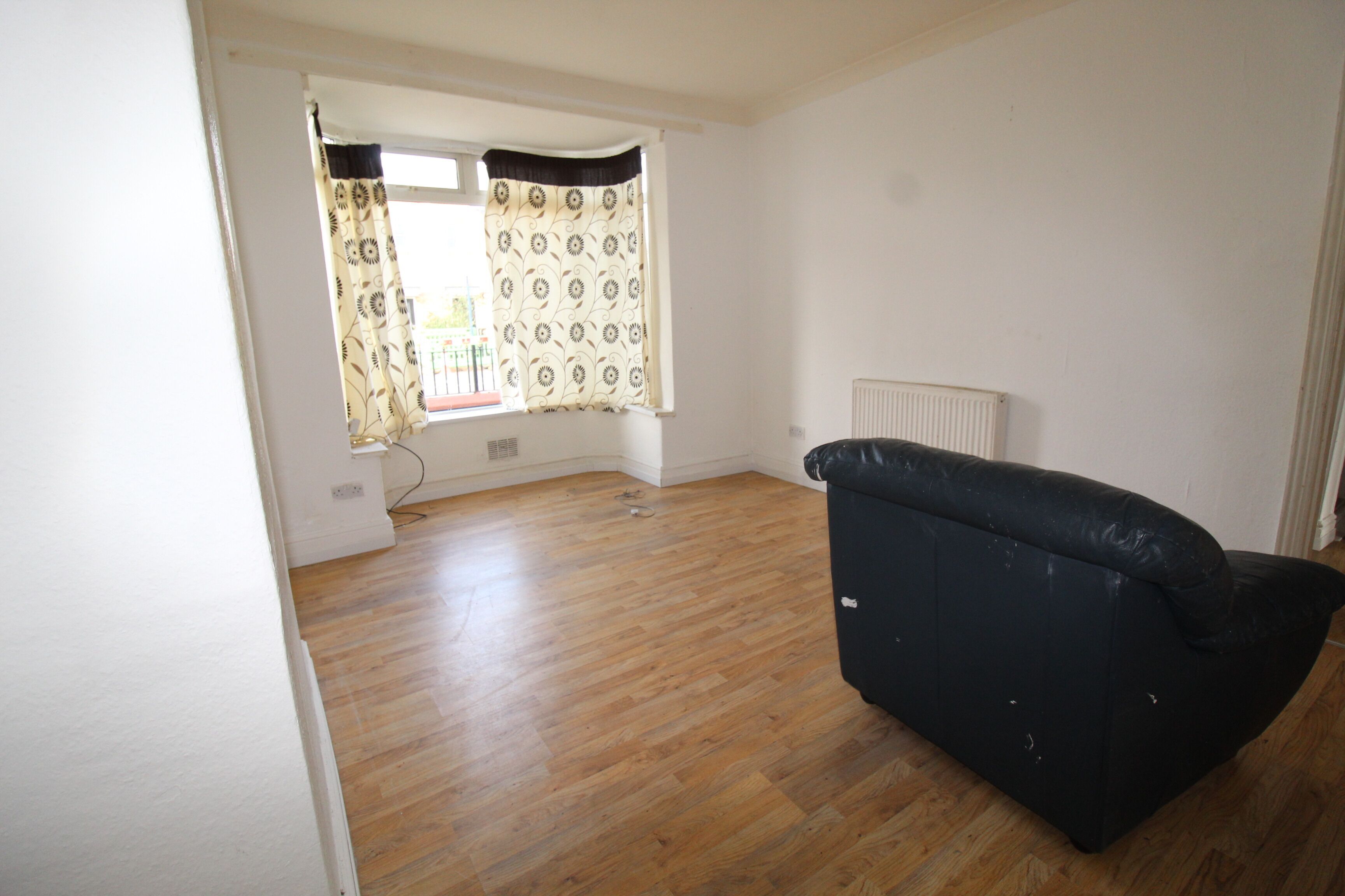 A room with wooden flooring, a black sofa, a radiator, and a window with patterned curtains.
