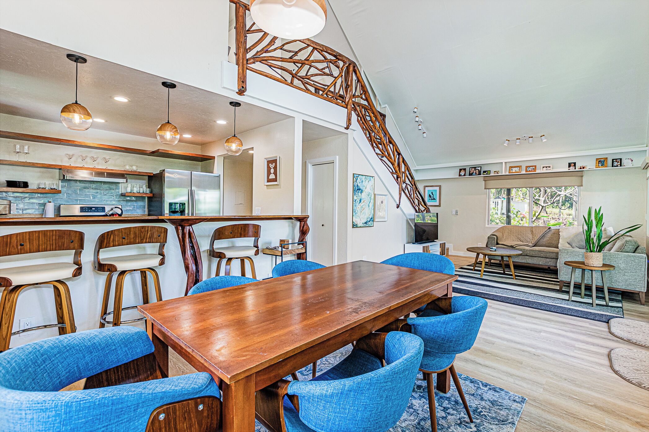 A spacious living area with a wooden dining table, blue upholstered chairs, a staircase, and a kitchen area with modern appliances.