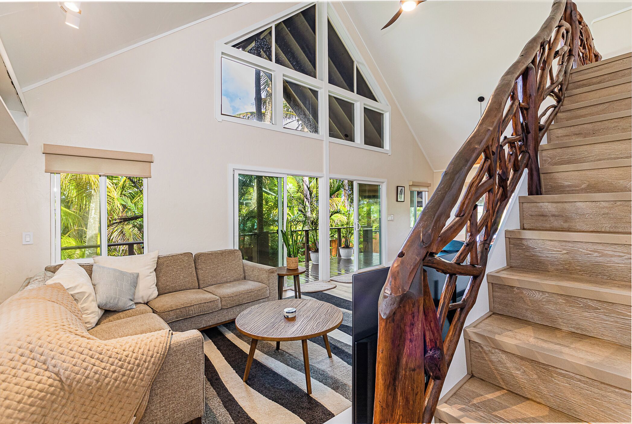 A spacious living area with a high ceiling, a wooden staircase, and large windows offering a view of greenery.