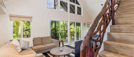 A spacious living area with a high ceiling, a wooden staircase, and large windows offering a view of greenery.