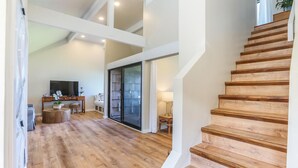A spacious living area with a wooden staircase, a flat-screen TV, and a sliding glass door leading to a balcony.