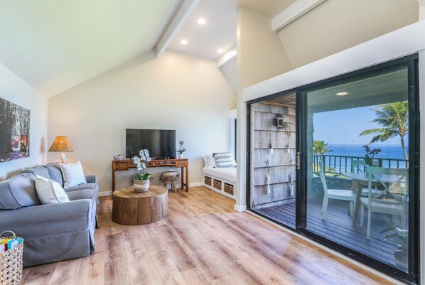 Una sala moderna con un sofá gris, una mesa de centro de madera y un televisor. Hay un balcón con vista al océano.