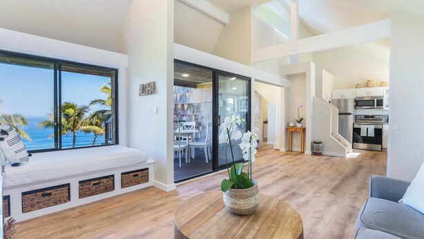 A modern living room with a large window overlooking the ocean, a seating area with a bench, a dining area, and a kitchen with stainless steel appliances.