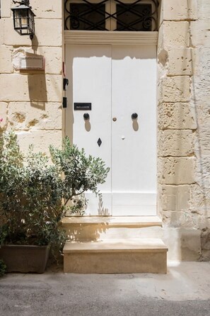 A white double door with a sign above it, a potted plant to the left, and a stone wall to the right. - Historic Maltese Townhouse (Rabat)
