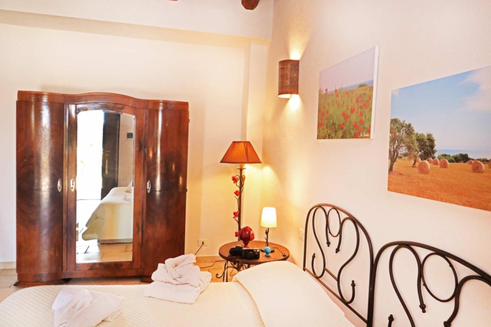 A bedroom with a bed, a wooden wardrobe, a bedside table with a lamp, and two paintings on the wall.