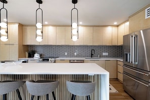 A modern kitchen with a central island, bar stools, stainless steel appliances, and pendant lighting.